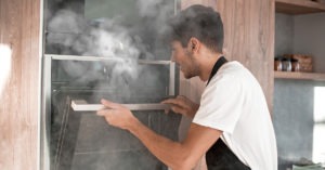 A person i an apron opening a defectice oven with smoke pouring out of it.