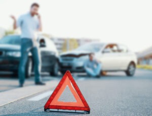 An orange triangle in the middle of the road with a man talking on the phone in front of crashed cars in the background.