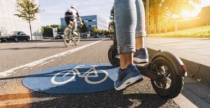 Person rides E-scooter on road next to car and bike.