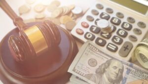 Table with calculator, judge gavel, and hundred dollar bills sit on table demonstrating legal damages.